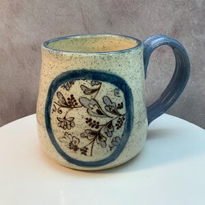 May include: A handmade ceramic mug with a speckled beige exterior and a blue handle. The mug features a blue circular design with floral and leaf patterns. The mug is approximately 4 inches tall.