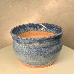May include: A round ceramic bowl with a wide opening and a curved body. The exterior features a mottled glaze in shades of deep blue, light blue, and white, while the interior and the base reveal a natural, speckled tan clay texture.