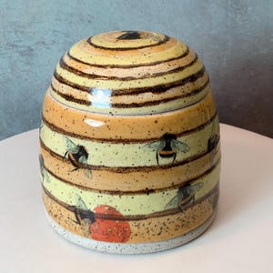 Puede incluir: Un tarro de cerámica con forma de colmena con tapa. El tarro presenta bandas horizontales alternas de color amarillo y naranja, con rayas marrones en la tapa. Ilustraciones de abejorros están dispersas alrededor del tarro. La base es blanca moteada.