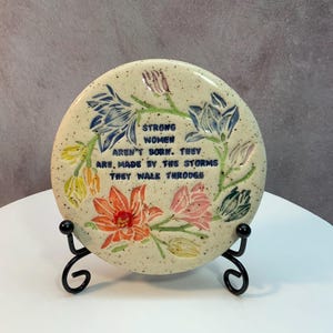 May include: A round, speckled ceramic plaque with floral designs in blue, orange, pink, and yellow. The plaque features the text "STRONG WOMEN AREN'T BORN. THEY ARE, MADE BY THE STORMS THEY WALK THROUGH." It is displayed on a black metal stand.