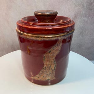 May include: A red ceramic jar with a lid and a knob. The jar is cylindrical with a flared rim. It has a brown and gold design on the side and a matching lid with a brown knob. The jar is likely for storage.