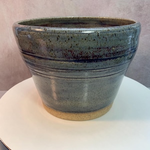 以下が含まれることがあります： 手作りの陶器のボウルで、幅が広く、わずかにフレアした形状をしています。ボウルは、濃い青と茶色のアクセントが付いた、斑点のある青灰色の釉薬が特徴です。ベースは釉薬がかかっておらず、自然なタン色の粘土が露出しています。ボウルの直径は約15cm、高さは約10cmです。