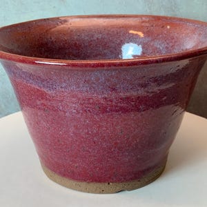May include: A handmade, glazed ceramic bowl with a wide, flared rim. The bowl features a rich, reddish-purple color with subtle variations in tone. The base is a natural, unglazed clay color. This decorative bowl is suitable for plants or as a decorative piece.