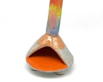 May include: A small ceramic incense burner with a rainbow-coloured chimney and a grey and orange base. The burner has three legs and a small opening for incense sticks.