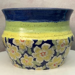 May include: A ceramic planter featuring a wide, flared rim with a deep blue glaze. The middle section transitions from a lime green to a bright yellow with dark speckles. The base displays a dark blue background with a pattern of yellow flowers.