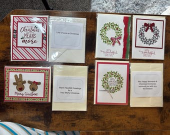 Puede incluir: Una colección de ocho tarjetas de Navidad con varios diseños. Las tarjetas presentan colores rojo y verde, coronas, renos y saludos navideños. El texto en las tarjetas incluye "Maybe Christmas means more", "Lots of Love at Christmas", "Warm Heartfelt Greetings for a Very Merry Christmas", "May Happy Moments & Cherished memories surround you with Joy this Season", y "Happy Wonderful Christmas".
