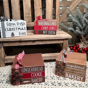 May include: Three stacked wooden boxes with Christmas-themed designs. The top box reads "Marshmallows Candy Canes Hot Cocoa", the middle box reads "Farm Fresh Christmas Trees", and the bottom box reads "Gingerbread Cookie Bakery".