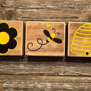 May include: Three wooden blocks with bee-themed designs. One block features a black and yellow flower, another a bee with a heart, and the last a yellow beehive. The blocks are arranged on a rustic wooden surface.