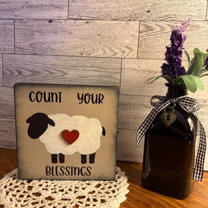 May include: A decorative wooden sign with the text "COUNT YOUR BLESSINGS" and a sheep illustration with a red heart. A black glass bottle with a gingham ribbon and purple flowers sits next to the sign. A white crocheted doily is under the sign.