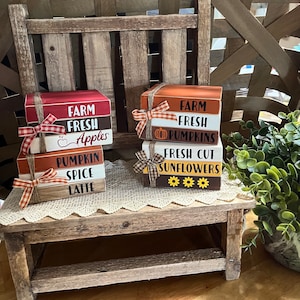May include: Two stacks of wooden blocks with fall-themed sayings. The first stack says "Farm Fresh Apples", "Pumpkin Spice Latte". The second stack says "Farm Fresh Pumpkins", "Fresh Cut Sunflowers". Both stacks are decorated with twine and plaid ribbon.