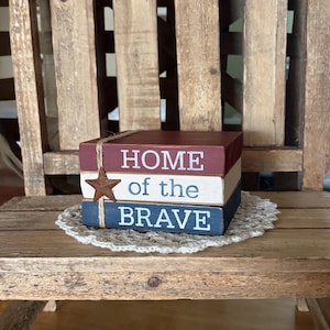May include: A decorative wooden block set with patriotic colors and the text "HOME of the BRAVE". The blocks are stacked and tied with twine, featuring a star accent. The set rests on a cream-colored doily, set against a rustic wooden background.