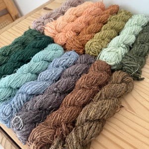 May include: A collection of yarn skeins in various colors, including shades of green, blue, brown, and peach. The yarn is arranged on a wooden surface, showcasing the natural tones and textures of the fiber. The skeins are tightly wound and ready for crafting.