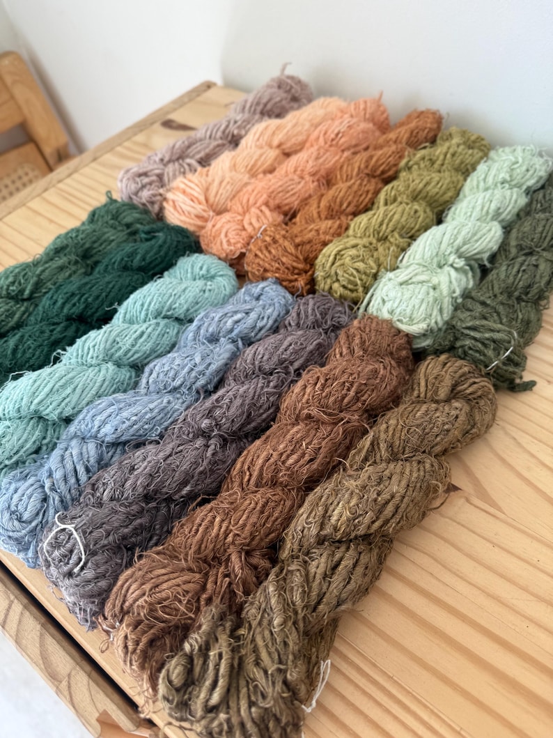May include: A collection of yarn skeins in various colors, including shades of green, blue, brown, and peach. The yarn is arranged on a wooden surface, showcasing the natural tones and textures of the fiber. The skeins are tightly wound and ready for crafting.