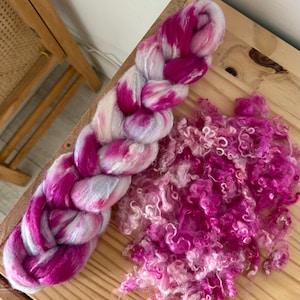 May include: A braided skein of pink and white yarn sits next to a pile of loose, curly pink fiber on a wooden surface. The yarn is a vibrant mix of colors, perfect for knitting or other fiber crafts.