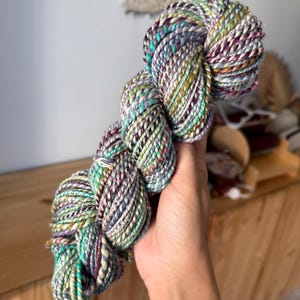 Puede incluir: Un ovillo de hilo colorido con una mezcla de tonos turquesa, morado y amarillo. El hilo est&aacute; torcido y parece suave, ideal para proyectos de tejido o ganchillo. La paleta de colores es vibrante y llamativa.