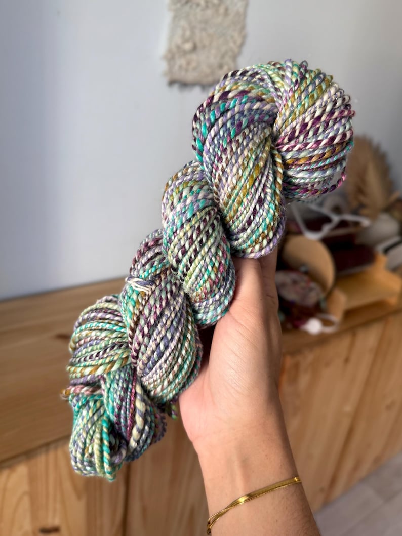 Puede incluir: Un ovillo de hilo colorido con una mezcla de tonos turquesa, morado y amarillo. El hilo est&aacute; torcido y parece suave, ideal para proyectos de tejido o ganchillo. La paleta de colores es vibrante y llamativa.