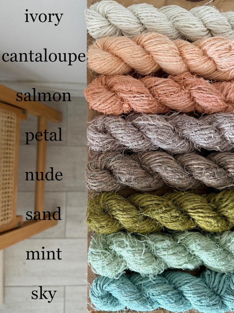 May include: A vertical display of eight skeins of yarn in various colors. The colors include ivory, cantaloupe, salmon, petal, nude, sand, mint, and sky blue. The yarn is tightly wound and appears to be made of a natural fiber.