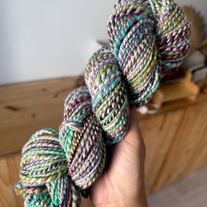 Puede incluir: Un ovillo de lana colorida con una mezcla de fibras turquesa, moradas, amarillas y blancas. La lana es gruesa y retorcida, con un aspecto texturizado. La lana se sostiene en una mano, mostrando sus colores vibrantes y su textura &uacute;nica, ideal para tejer o hacer ganchillo.