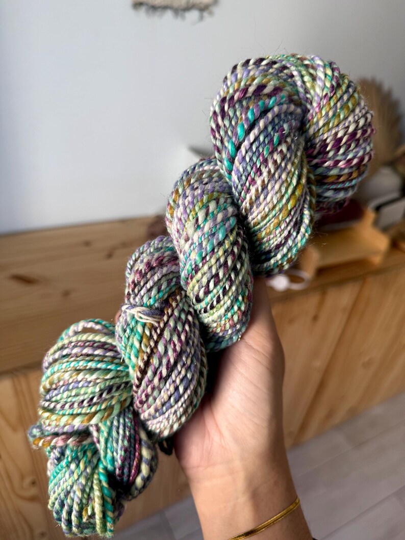 Puede incluir: Un ovillo de lana colorida con una mezcla de fibras turquesa, moradas, amarillas y blancas. La lana es gruesa y retorcida, con un aspecto texturizado. La lana se sostiene en una mano, mostrando sus colores vibrantes y su textura &uacute;nica, ideal para tejer o hacer ganchillo.
