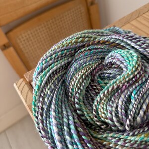 Puede incluir: Un ovillo de lana colorida en tonos turquesa, morado, verde y blanco. La lana est&aacute; bien enrollada y descansa sobre una superficie de madera, con una silla de madera en el fondo. La lana parece ser hilada a mano o artesanal.