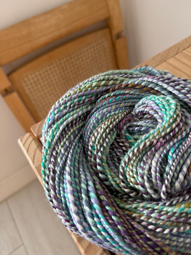 Puede incluir: Un ovillo de lana colorida en tonos turquesa, morado, verde y blanco. La lana est&aacute; bien enrollada y descansa sobre una superficie de madera, con una silla de madera en el fondo. La lana parece ser hilada a mano o artesanal.