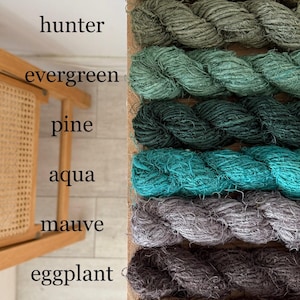 May include: A display of colorful yarn in shades of khaki, hunter green, evergreen, pine, aqua, mauve, eggplant, and chocolate. The yarn is arranged in thick, twisted ropes, with the color names written above each. The yarn is displayed on a wooden board.