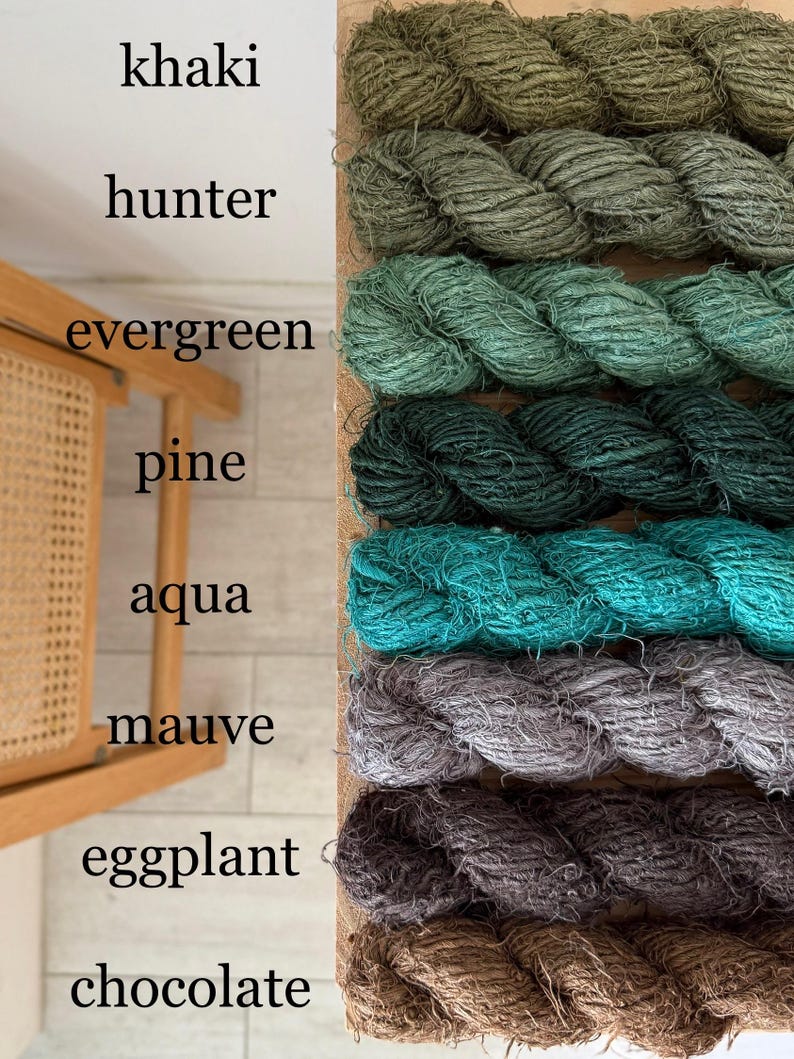 May include: A display of colorful yarn in shades of khaki, hunter green, evergreen, pine, aqua, mauve, eggplant, and chocolate. The yarn is arranged in thick, twisted ropes, with the color names written above each. The yarn is displayed on a wooden board.