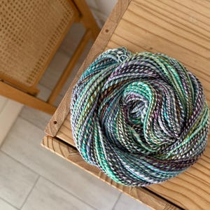 Puede incluir: Una madeja de hilo multicolor, principalmente turquesa, morado, verde y blanco, descansa sobre una superficie de madera. El hilo parece estar te&ntilde;ido a mano, con sutiles variaciones de color. La textura del hilo es visible.