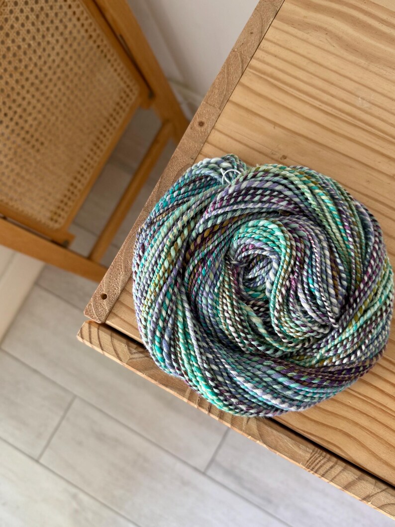 Puede incluir: Una madeja de hilo multicolor, principalmente turquesa, morado, verde y blanco, descansa sobre una superficie de madera. El hilo parece estar te&ntilde;ido a mano, con sutiles variaciones de color. La textura del hilo es visible.