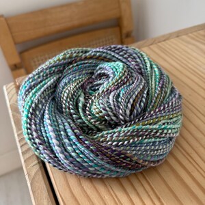 Puede incluir: Una madeja de lana grande y bien enrollada en tonos turquesa, morado, blanco y marr&oacute;n. La lana est&aacute; sobre una superficie de madera clara. La lana parece estar te&ntilde;ida a mano y es adecuada para proyectos de tejido o ganchillo.