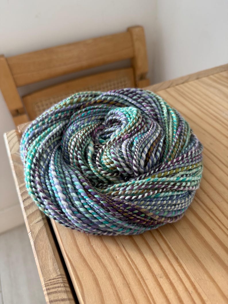 Puede incluir: Una madeja de lana grande y bien enrollada en tonos turquesa, morado, blanco y marr&oacute;n. La lana est&aacute; sobre una superficie de madera clara. La lana parece estar te&ntilde;ida a mano y es adecuada para proyectos de tejido o ganchillo.