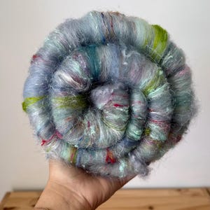 May include: A circular coil of fluffy, multi-colored fiber roving featuring a blend of soft blue, grey, and teal tones with vibrant accents of lime green, magenta, and white. The texture is fuzzy and fibrous, arranged in a spiral pattern.