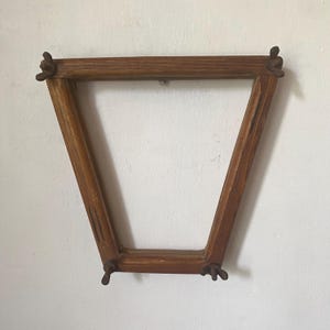 Peut inclure: Un cadre en bois en forme de V, avec une finition naturelle veinée. Le cadre comporte des éléments décoratifs à chaque coin. Le cadre est accroché à un mur blanc.