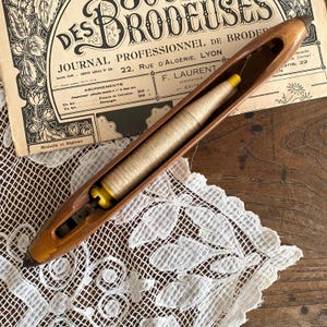 Può includere: Una spola di legno con una bobina di filo gialla all'interno, appoggiata su un centrino di pizzo bianco. La spola è accanto a una rivista francese vintage intitolata "DES BRODEUSES" con testo in francese.