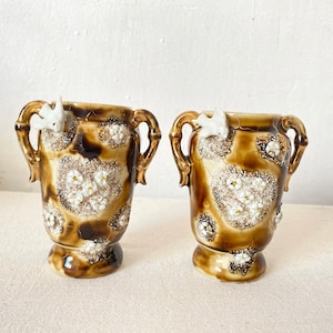 Op de afbeelding: Twee kleine, bruine keramische vazen met witte bloemenmotieven en gouden accenten. Elke vaas heeft een vogelbeeldje op de handgreep.