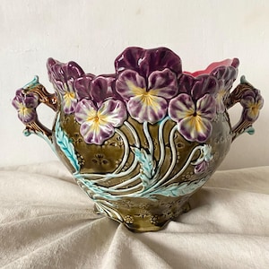 Può includere: Una ciotola decorativa in ceramica con un motivo floreale. La ciotola presenta viole del pensiero viola in rilievo con centri gialli, foglie verdi e accenti marroni. Le maniglie sono marroni con piccoli fiori viola.