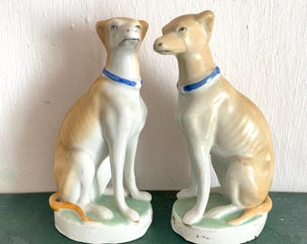May include: Two porcelain figurines of greyhounds sitting on green bases. The greyhounds are white with brown markings and have blue collars.