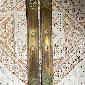 Pode incluir: Duas placas de dedo de porta antigas em latão. Cada placa é retangular com desenhos florais e geométricos ornamentados. As placas têm buracos de fechadura e aberturas circulares. O latão tem uma pátina quente e envelhecida.