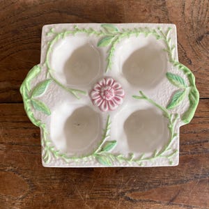 Può includere: Un portauova in ceramica bianca con quattro rientranze circolari. Il supporto è decorato con foglie e tralci verdi sui bordi e un fiore rosa al centro. Il supporto è su una superficie di legno.