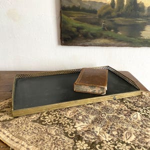 Puede incluir: Una bandeja rectangular negra con un marco dorado y un borde de celosía decorativo. Un pequeño libro encuadernado en cuero marrón y desgastado descansa sobre la bandeja. La bandeja está sobre una superficie estampada, con una pintura al fondo.