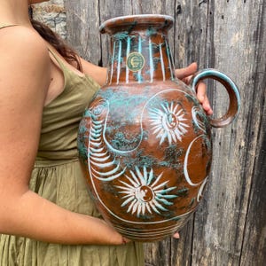 Op de afbeelding: Een grote, bruine keramische kruik met een handvat, versierd met witte en turquoise ontwerpen. De kruik heeft een brede basis, een smalle hals en een groot handvat. Het ontwerp omvat zonachtige vormen en gebogen lijnen.