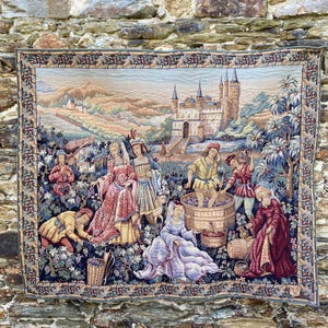 May include: A rectangular woven tapestry featuring a medieval grape harvest scene with figures in period clothing, a large wooden barrel, and a stone castle in the background. The border displays a repeating floral pattern in blue and gold tones.