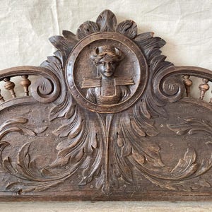 Può includere: Testata in legno antico con intagli intricati. Il design include un medaglione centrale con un busto di donna, circondato da motivi floreali e fogliari. Il legno è di un marrone scuro, che mette in risalto l'età e l'artigianato.