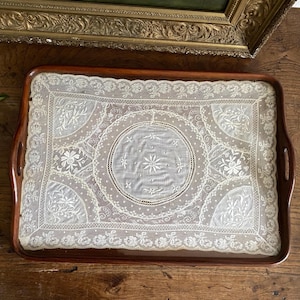 May include: A rectangular wooden serving tray with a dark brown finish. The tray features a delicate white lace insert with floral and circular patterns. The tray has integrated handles on each side, offering a classic and elegant design.