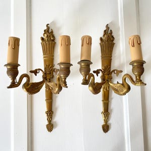 May include: Two antique brass wall sconces with swan and flame details. Each sconce holds two candle-shaped light bulbs. The sconces are mounted on a white wall. The sconces have a vintage, ornate design.