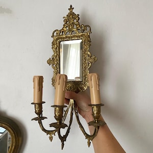 Könnte beinhalten: Verzierte goldfarbene Wandleuchte mit einem kleinen rechteckigen Spiegel und drei Kerzenhaltern. Die Leuchte weist detaillierte Schnörkel und Engel-Akzente auf. Die Kerzen sind hellbeige und verleihen eine Vintage-Ästhetik.