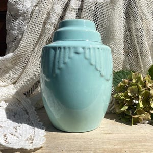 Puede incluir: Un tarro de cerámica azul claro con una tapa escalonada y un patrón decorativo en forma de goteo alrededor del hombro superior. La superficie esmaltada presenta una fina red de líneas de craquelado por todo el cuerpo.