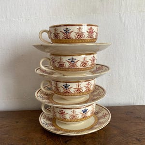 Könnte beinhalten: Ein Stapel aus vier cremefarbenen Keramiktassen und Untertassen, die jeweils mit einem wiederkehrenden Muster aus stilisierten roten und blauen Blumenmotiven und goldenen geometrischen Rändern verziert sind. Die Tassen sind rund geformt.