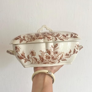 Può includere: Piatto da portata color crema con coperchio e motivo floreale marrone. Il piatto ha un bordo smerlato e due manici. Il coperchio presenta una maniglia decorativa. Si vede il polso di una persona con un braccialetto.