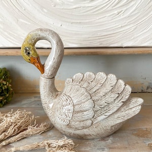 May include: A ceramic swan figurine with a glossy off-white finish and textured feather details. The swan has a yellow head, a bright orange beak, and painted black eyes with eyelashes. The neck is curved, and the body is shaped like a lidded container.